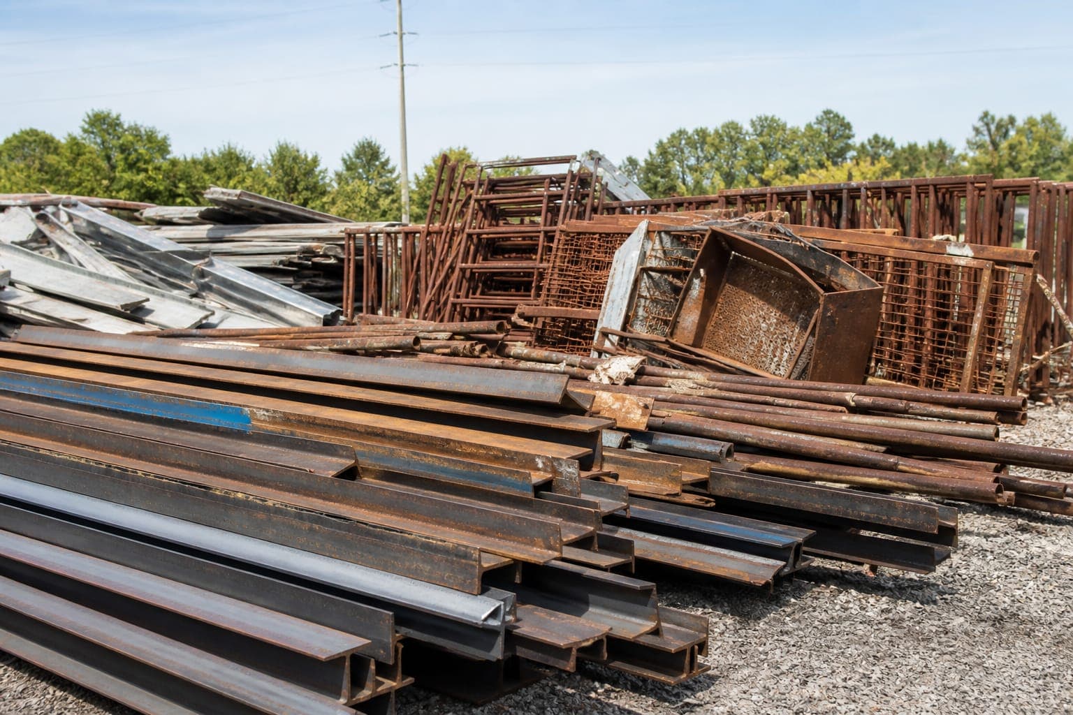 Stacked steel I-beams and rusted farm iron in an outdoor yard.